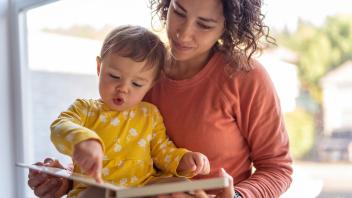 toddler pointing to words in a picture book while sitting on mother's lap