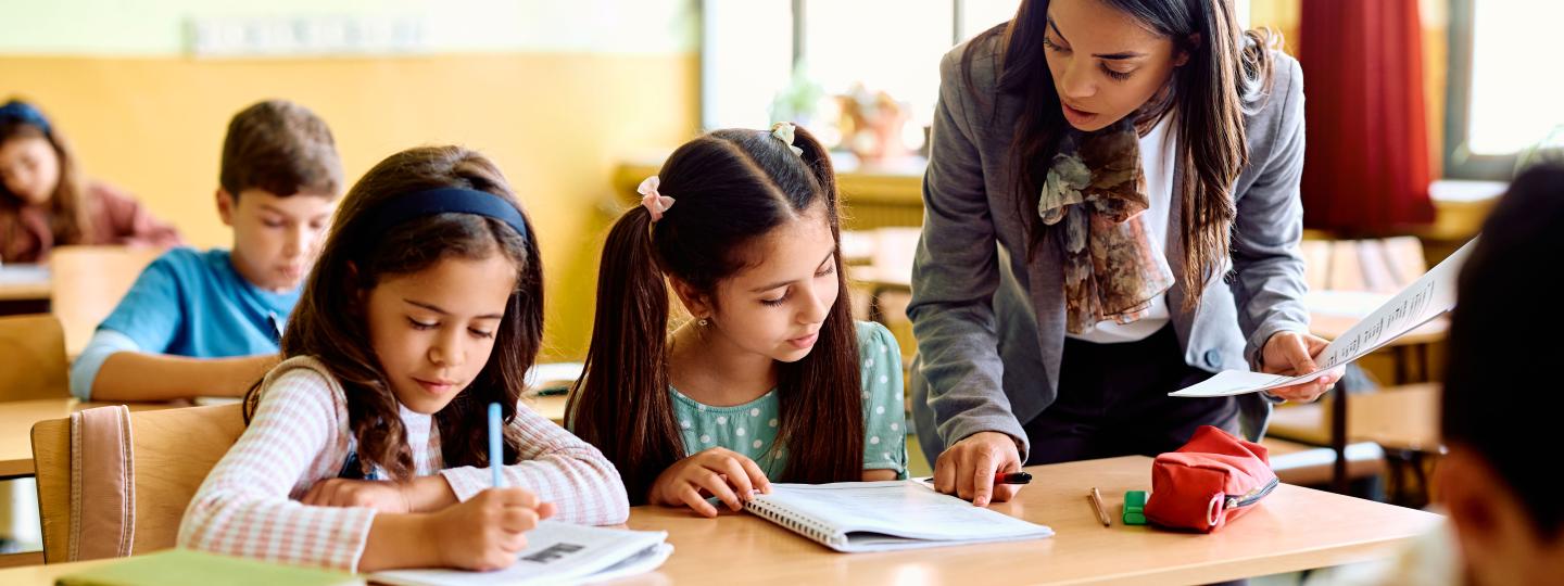 elementary teachers talking with two students at their desks